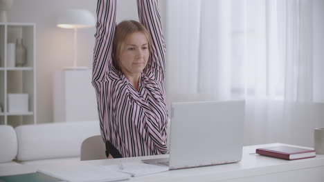 student-woman-is-learning-online-with-laptop-and-copybook-at-home-stretching-tired-muscles-of-back-and-neck
